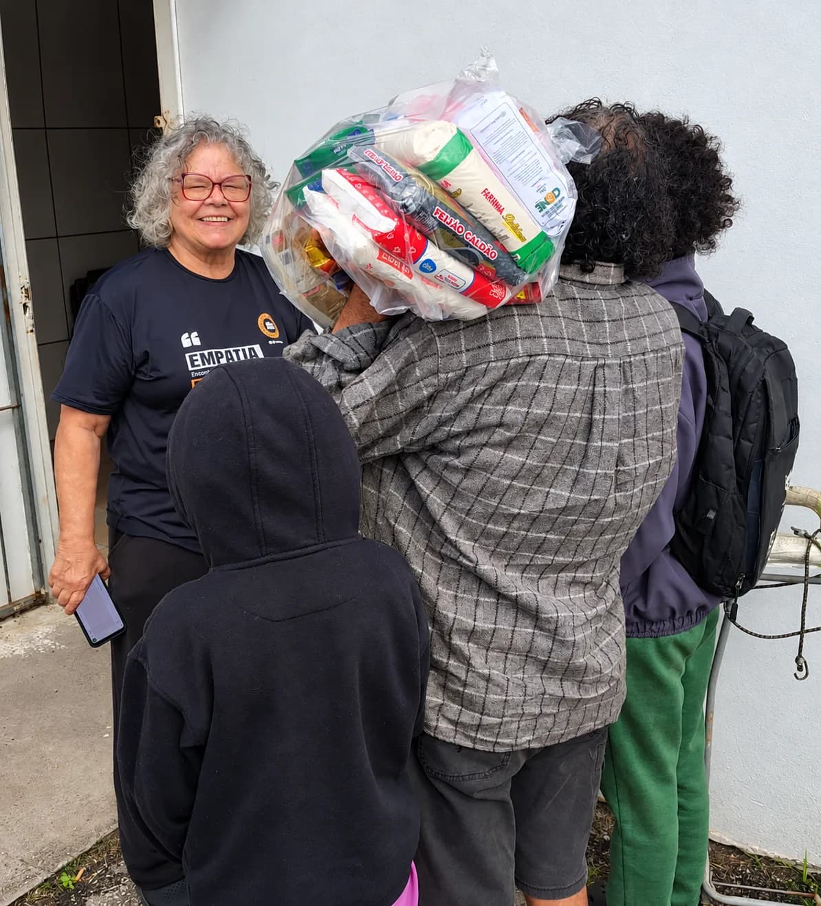 Voluntária do Nós na Rua sorrindo ao lado de uma família, de costas, recebendo uma cesta básica na porta de casa.